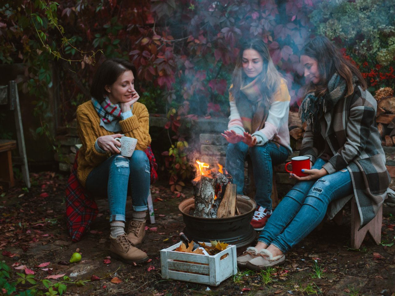 Three women sitting around a backyard fire pit with mugs, talking easily