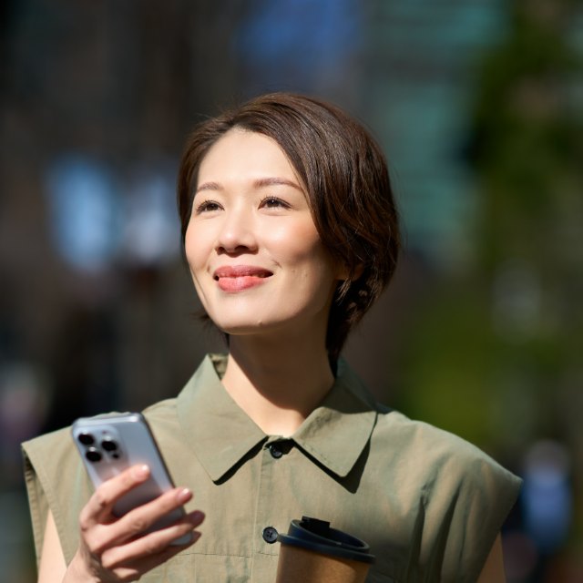 A woman walking with coffee and her phone, smiling into the sun